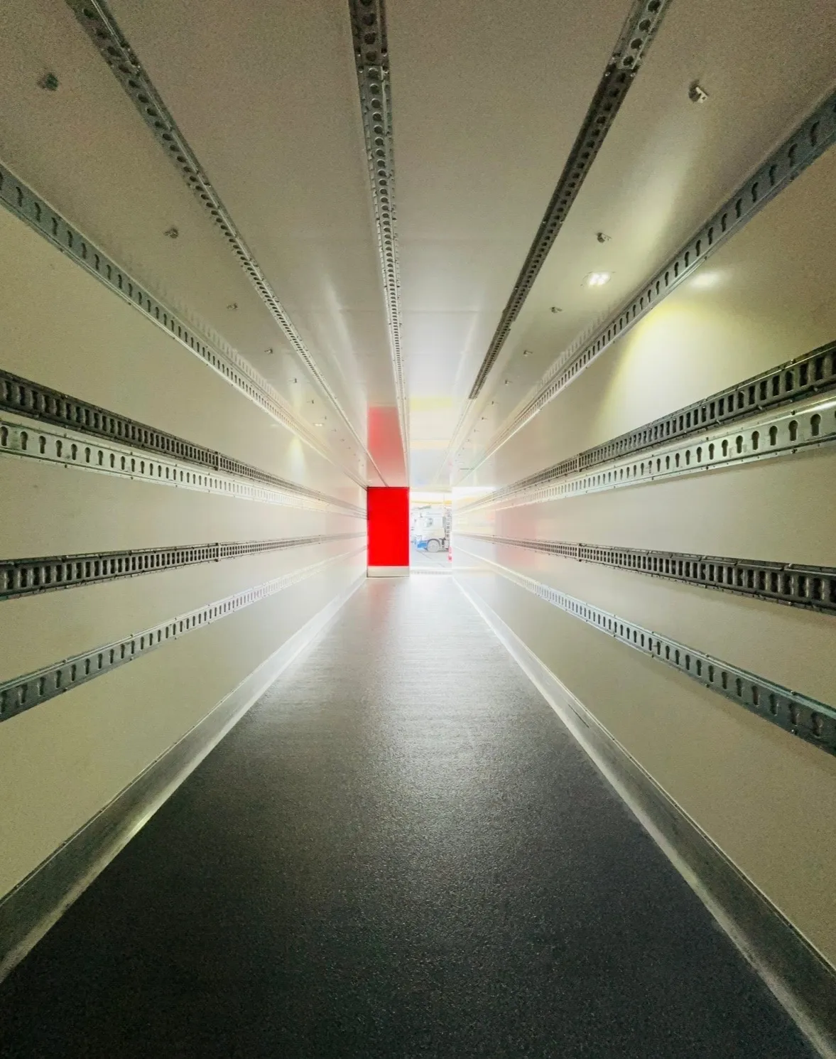 Blik door een lege, kraaknette Malatra-trailer, met een rode deur die licht binnenlaat op het einde